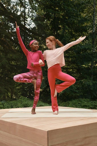 Two young girls in yoga poses on a wooden platform surrounded by trees, wearing pink athletic clothing and smiling at each other outdoors.