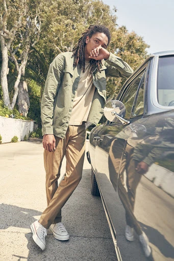 A young adult male with dreadlocks poses confidently, leaning against a vintage car in olive shirt and khaki pants on a sunny day.