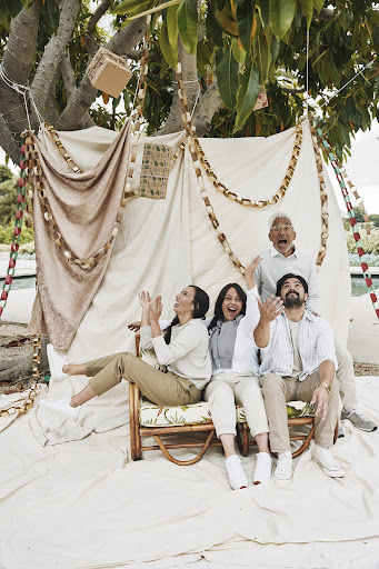 A family of five gathers in front of a tent with a beige tarp and decorative lights, all smiling with arms up in an outdoor setting.