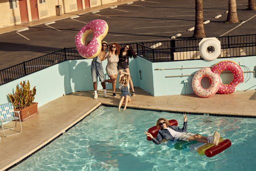 Group of friends enjoy a sunny day at a pool with donut-shaped floats in a vintage-style motel setting, capturing a fun, summery vibe.