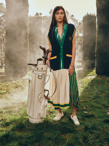 Adult female poses outdoors wearing sleeveless cardigan with a golf bag on a grassy lawn, trees and some fog in the background.