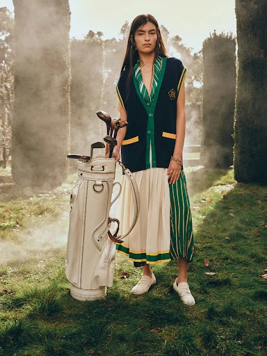 Adult female poses outdoors wearing sleeveless cardigan with a golf bag on a grassy lawn, trees and some fog in the background.