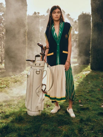 Adult female poses outdoors wearing sleeveless cardigan with a golf bag on a grassy lawn, trees and some fog in the background.