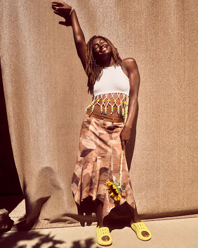 Woman in a white top and patterned skirt with sunflower stands in front of a textured brown background, smiling with her arm raised.