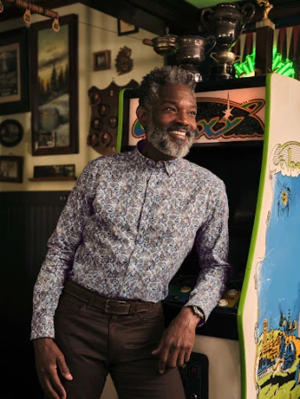 A smiling Black man in a patterned shirt stands indoors next to a vintage arcade game, creating a nostalgic and inviting scene.