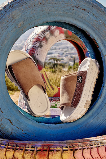 A pair of brown slide sandals displayed inside a blue-painted tire against an out-of-focus urban background on a sunny day.
