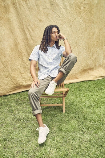 An adult male with dreadlocks is sitting on a wooden chair in a grassy area with a neutral-toned backdrop, wearing a patterned shirt and khaki pants.