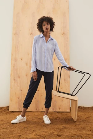 Woman with curly dark hair wearing a blue striped button-down shirt and dark trousers standing on dirt before a large plywood panel.