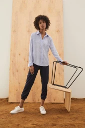 Woman with curly dark hair wearing a blue striped button-down shirt and dark trousers standing on dirt before a large plywood panel.