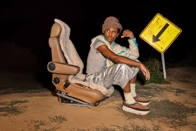 Young man in chunky knit beanie and shiny trousers sitting on a car seat outdoors at night next to a yellow arrow sign