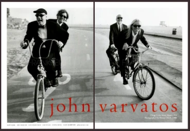 Black and white editorial photograph of two men in suits riding a tandem bicycle on a paved coastal path.