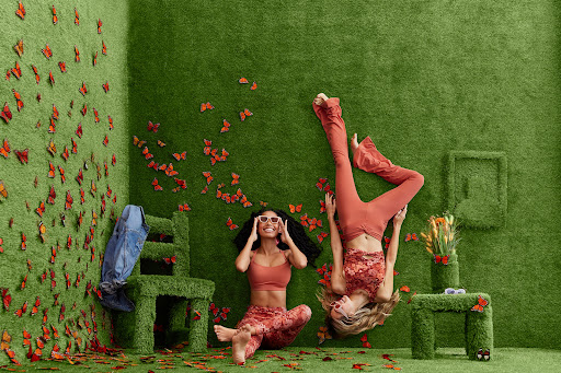 Two women in orange outfits pose in a vibrant green room adorned with butterflies, seated near a chair, table and frame covered in grass.