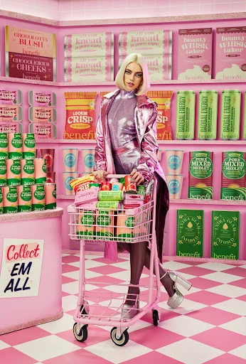 Woman in shiny jacket poses with a shopping cart full of beauty products in an all-pink store with Benefit Cosmetics merchandise.