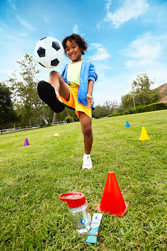 Young child in motion, playfully kicking a soccer ball on a vibrant green field under a blue sky, with sports cones nearby.