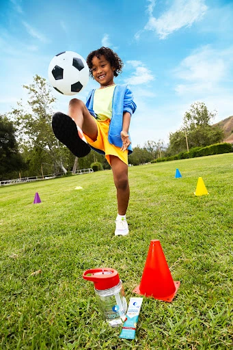 Young child in motion, playfully kicking a soccer ball on a vibrant green field under a blue sky, with sports cones nearby.