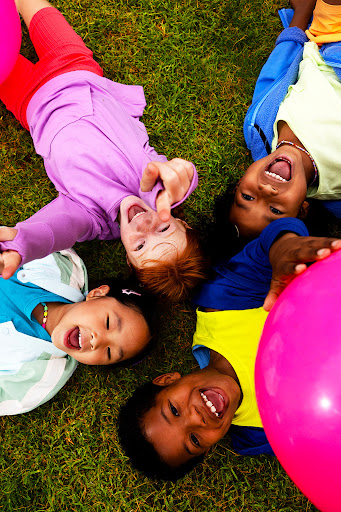 Overhead view of four smiling multiethnic children lying on green grass with a pink balloon, facing the camera with open mouths.