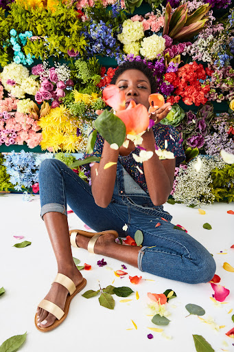 A woman with dark hair, wearing overalls and sandals, is blowing pink and white flower petals into the air in a colorful floral setting.