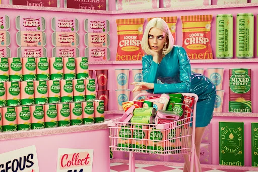 Woman poses with Benefit Cosmetics products in a whimsical, pastel-colored shopping scene featuring a pink shopping cart and product displays.