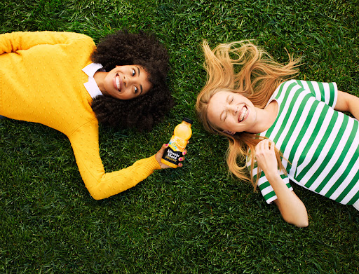 Two young women smile lying on green grass with a bottle of Minute Maid juice in bright, natural light. Top-down perspective.