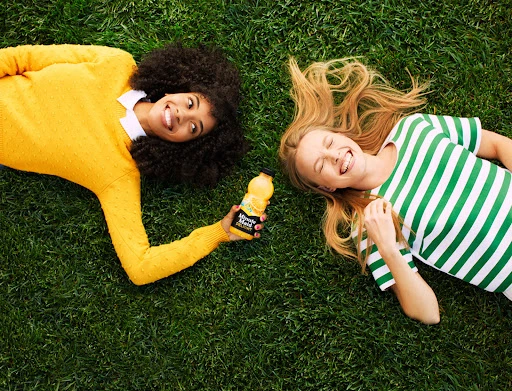 Two young women smile lying on green grass with a bottle of Minute Maid juice in bright, natural light. Top-down perspective.