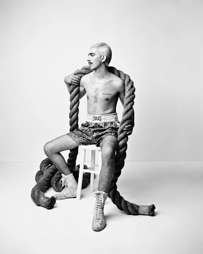 Black and white studio shot of a shirtless adult male boxer sitting on a stool with a thick rope draped around his neck and shoulders.
