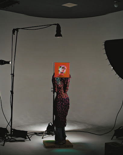 Fashion photography of a figure in a textured dress holding a portrait in a photo studio setting with lighting and backdrop equipment.