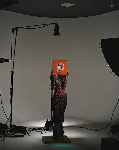 Fashion photography of a figure in a textured dress holding a portrait in a photo studio setting with lighting and backdrop equipment.