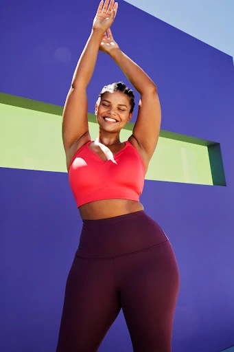 A smiling adult female poses in athletic wear in front of a colorful wall, raising her arms in an outdoor setting during daylight.