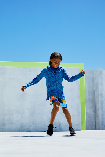 Enthusiastic child with braids dances in a blue hooded jacket and colorful shorts against a sunny sky and modern architectural backdrop.