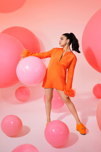 Woman in orange dress with feathered sleeves poses with pink balloons in a photo studio, wearing matching orange platform shoes.