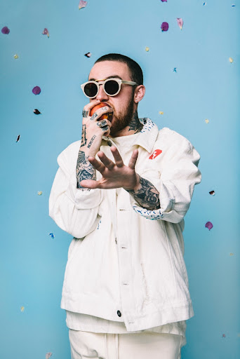Portrait of a tatted male wearing sunglasses, a white jacket, and eating an orange against a blue background with confetti.