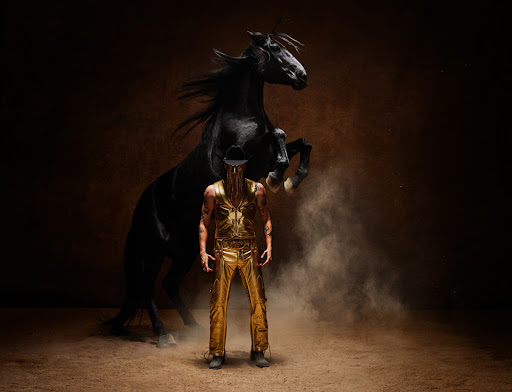 Dramatic studio portrait of a muscular man in a gold costume standing in front of a rearing black horse against a dark brown background.