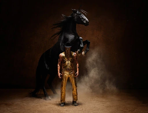 Dramatic studio portrait of a muscular man in a gold costume standing in front of a rearing black horse against a dark brown background.