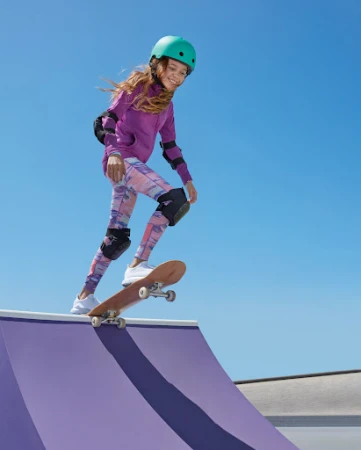 Young female skateboarder wearing a purple hoodie, turquoise helmet, and protective gear riding down a ramp