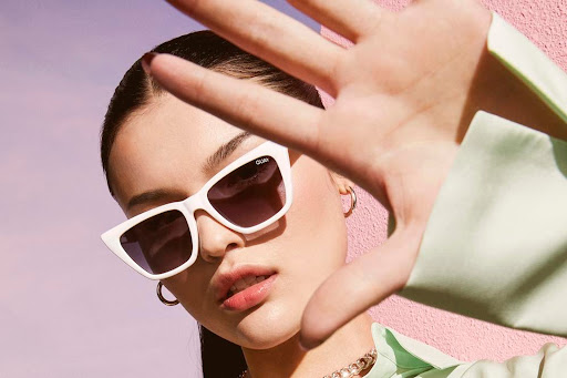 Close-up of a stylish woman wearing trendy white sunglasses and a mint green top, posing against a pink wall in an outdoor setting.