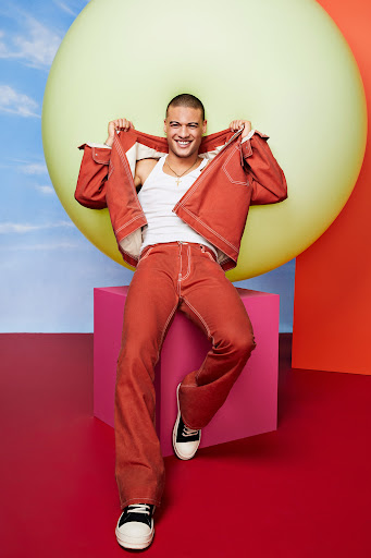 Smiling man poses indoors wearing a red denim jacket, white top, and red jeans, set against colorful geometric shapes and balloons.