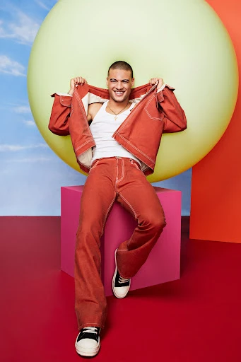 Smiling man poses indoors wearing a red denim jacket, white top, and red jeans, set against colorful geometric shapes and balloons.
