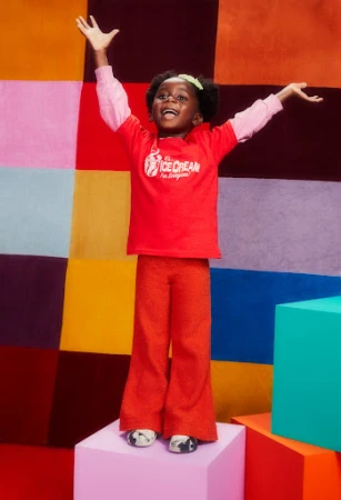 A young child with arms raised poses on a pink block in a red top and textured orange pants against a colorful geometric backdrop.