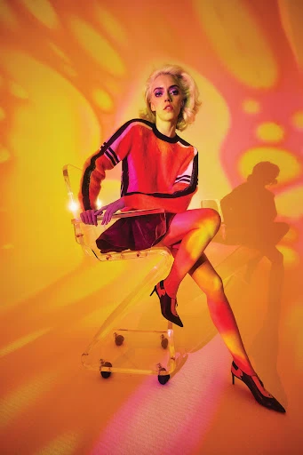 A fashionable blonde woman poses in a transparent chair under orange and pink lighting wearing a colorful top and heels indoors.