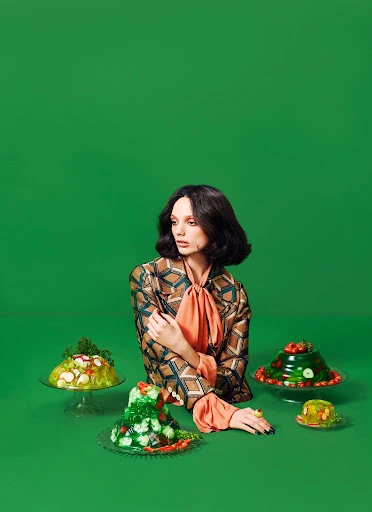 Portrait of a woman in a patterned jacket and orange bow, surrounded by aspic food on glass stands against a green backdrop.