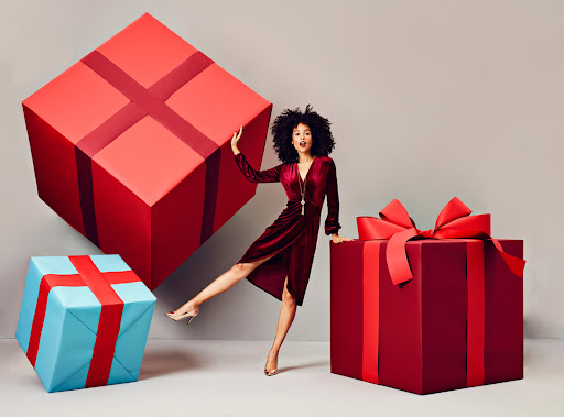 Woman in a velvet dress striking a pose next to oversized wrapped gifts against a neutral backdrop, creating a festive holiday atmosphere.
