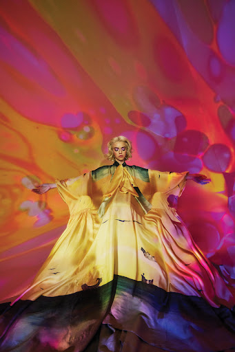 Adult woman wearing a long yellow and green dress stands with her arms outstretched against a vibrant background with orange and purple tones.