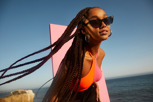A woman with braids and sunglasses poses outside near the ocean. She is wearing a colorful halter top and gold hoop earrings.