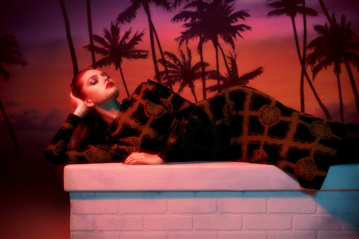 Adult female fashion model in patterned outfit rests against a white wall with palm trees and an orange-pink sky backdrop.