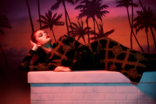 Adult female fashion model in patterned outfit rests against a white wall with palm trees and an orange-pink sky backdrop.