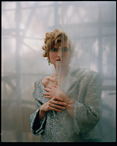 Woman with retro hair and makeup wearing an embellished jacket poses in front of a white translucent backdrop in a medium studio shot.