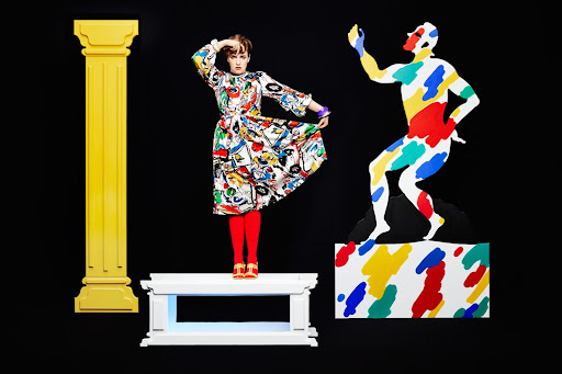Woman in patterned dress poses with colorful cutout figure and yellow column on a platform in a brightly lit photo studio.