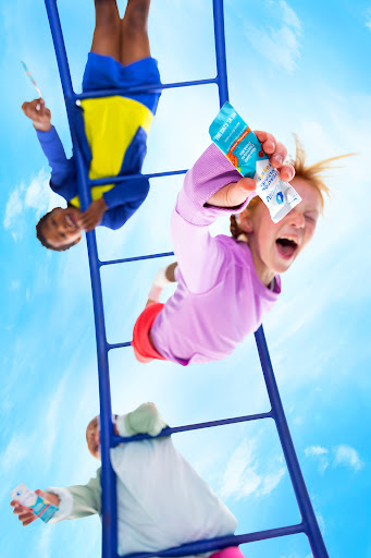 Three children climbing blue jungle gym, smiling, and holding snack pouches against a bright blue sky filled with fluffy white clouds.