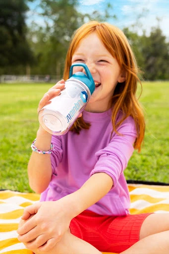 A happy red-haired child drinks from a clear water bottle with blue accents while sitting on a striped blanket in a grassy outdoor setting.