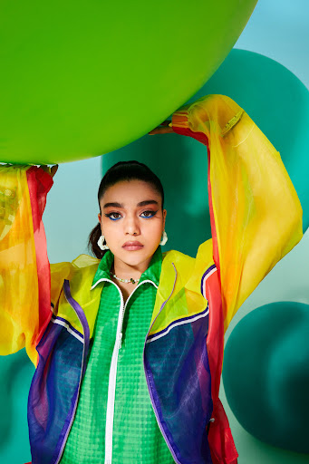 A young adult woman poses with a large green balloon, wearing a colorful sheer jacket, against a light blue backdrop with green balloons.